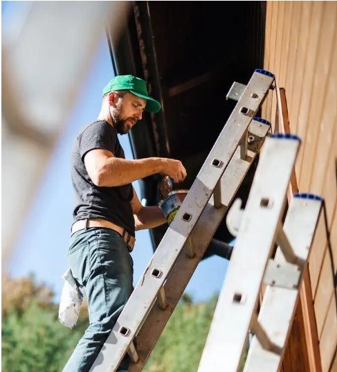 A Next Level painter on a ladder cutting in exterior trim under a clear sky